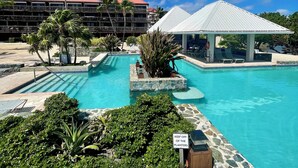 A resort pool area with a clear blue sky, palm trees, and a building with a red roof. - Sapphire Beach, Beach Level Condo, B Bldg, Bella Mar, Remodeled 2025, Generator (St. Thomas)
