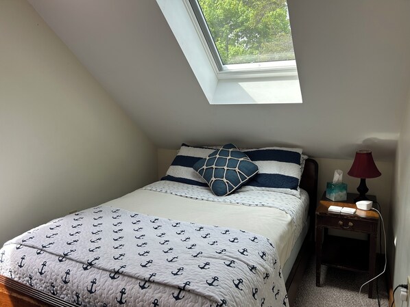 A bedroom with a sloped ceiling, a skylight, a bed with a patterned quilt, pillows, a bedside table with a lamp, and a tissue box.