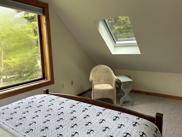 A bedroom with a sloped ceiling, a bed with a white quilt, a wicker chair, a small table, and a skylight overlooking greenery. - Peaceful Cottage with Ocean Access (Southwest Harbor)