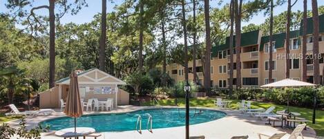 A hotel with a swimming pool surrounded by lounge chairs and a small building with a sign.
