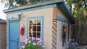 A small, single-story building with a blue door and window, surrounded by a garden with various flowers. - Casa Madre 3 bedroom/2 baths (Taos)