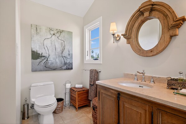 A bathroom with a white toilet, a wooden vanity with a sink, a large mirror, and a painting on the wall.