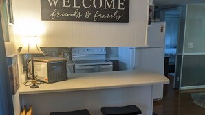 Une cuisine avec une pancarte indiquant « BIENVENUE, amis et famille », une cuisinière, un four et un réfrigérateur. On y trouve également deux tabourets et une lampe.