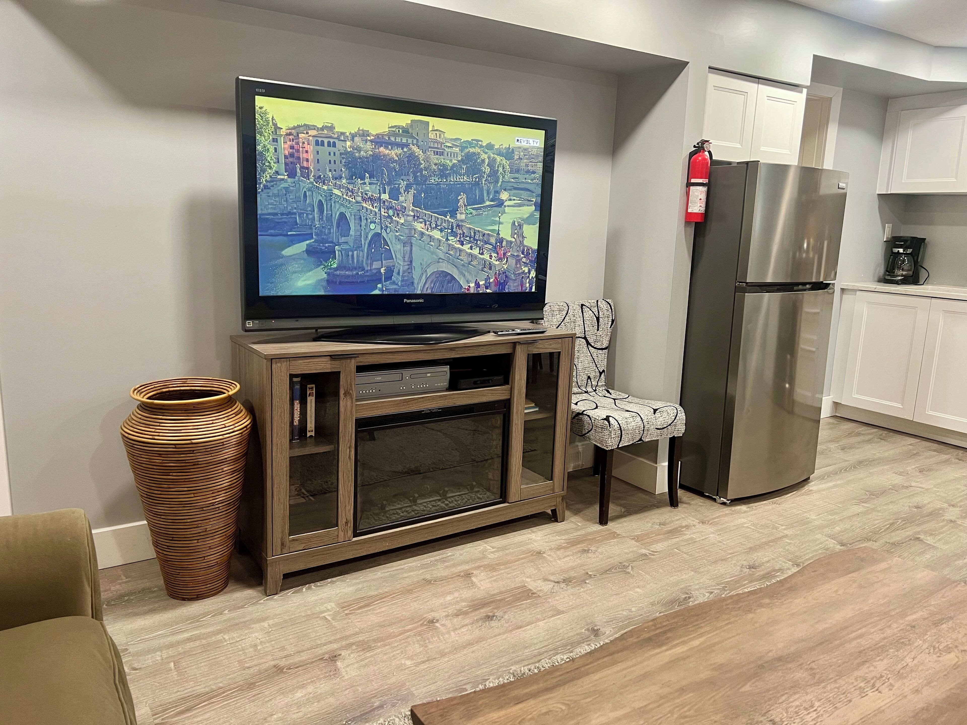 A living room with a television on a wooden stand, a wicker vase, a patterned chair, a kitchen area with a refrigerator, and a fire extinguisher on the wall.