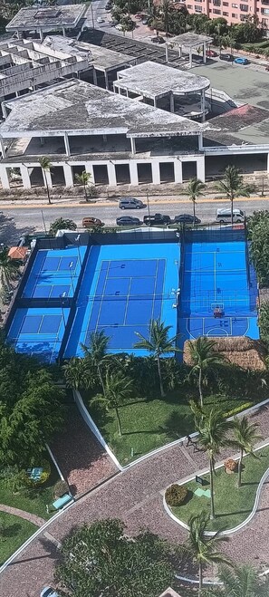 Vue aérienne d’un quartier résidentiel avec un grand terrain de tennis bleu, entouré de palmiers et d’un parking.