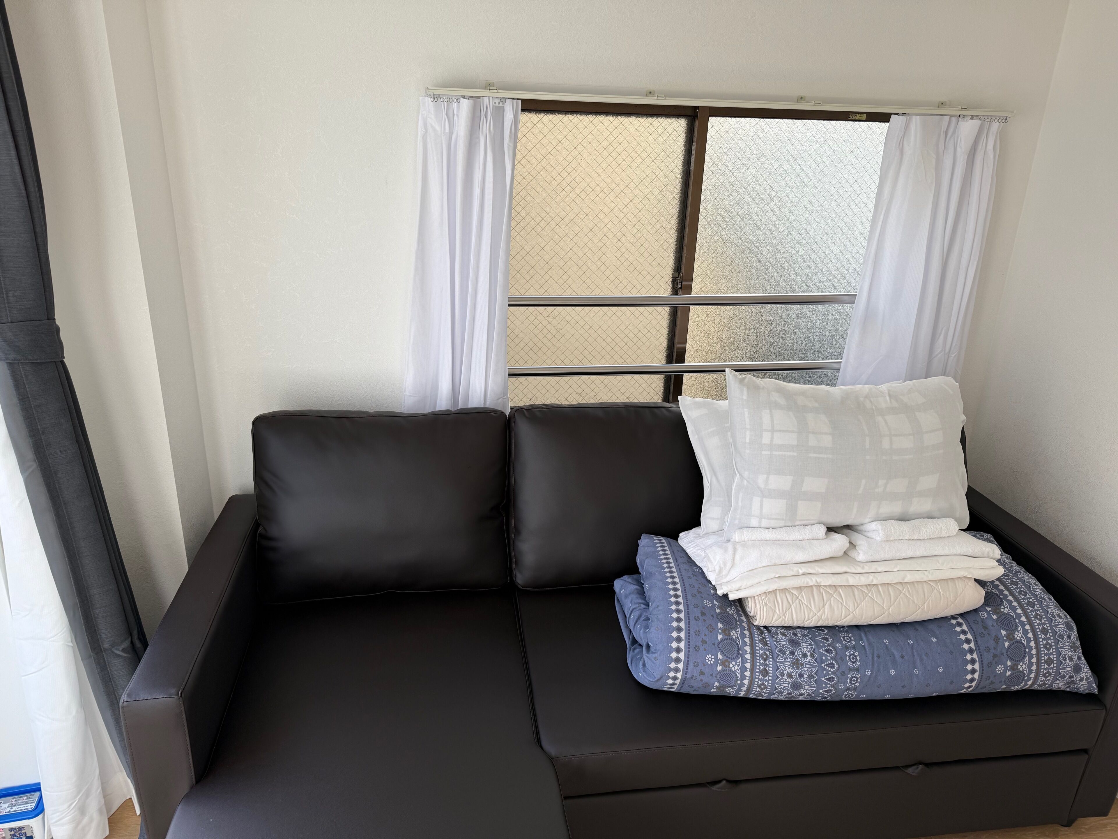 A black sofa with a patterned pillow and folded blankets, a window with white curtains, and a metal railing.