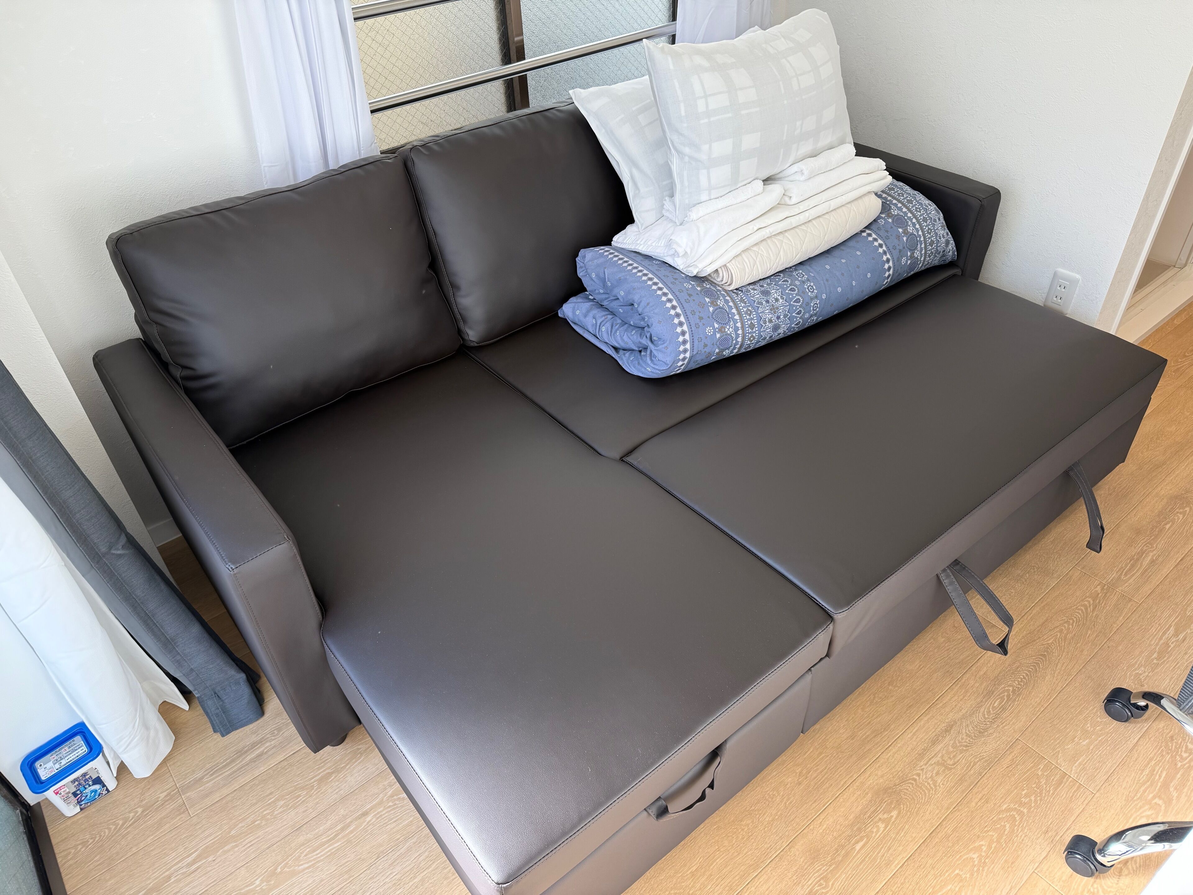 En svart soffa med ett utdragbart bord, ett fönster med utsikt och en liten blå papperskorg.