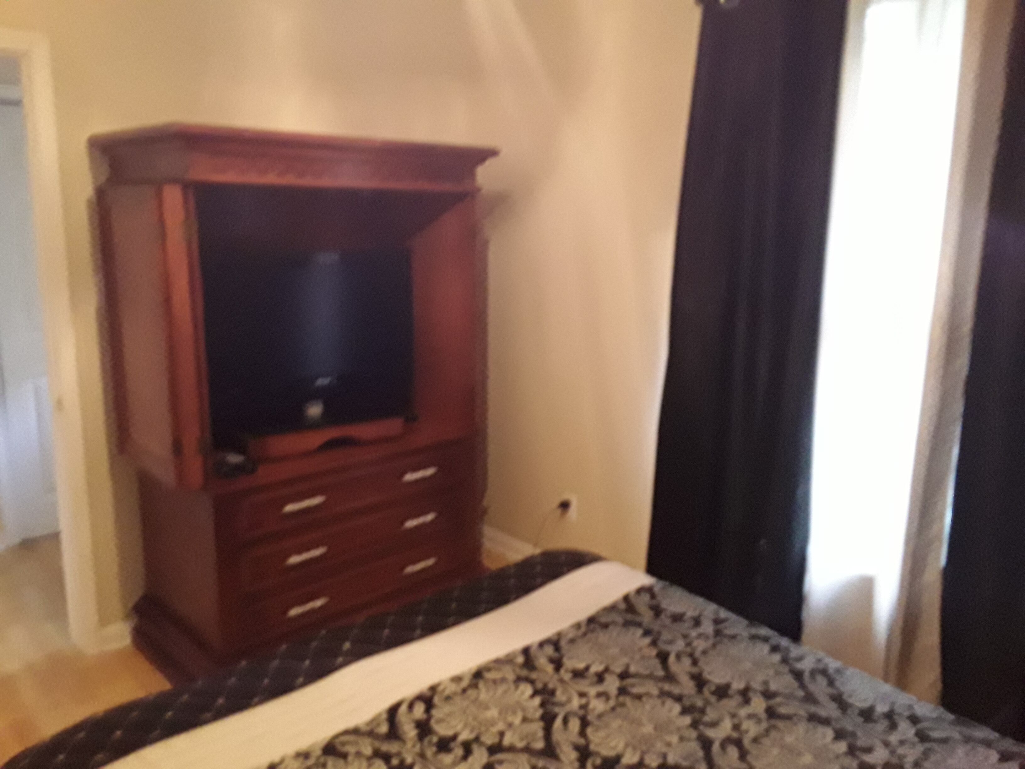 Ein Schlafzimmer mit einem hölzernen Schminktisch, einem Bett mit gemustertem Bettbezug und einem Fenster mit Vorhängen.