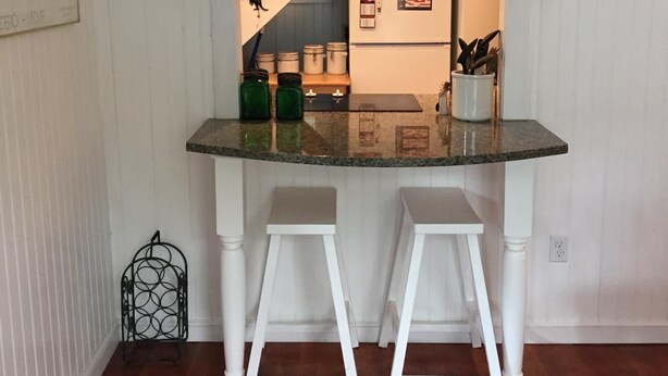 Una cocina con una encimera de isla de vidrio, taburetes blancos, una lámpara colgante y un refrigerador con imanes.