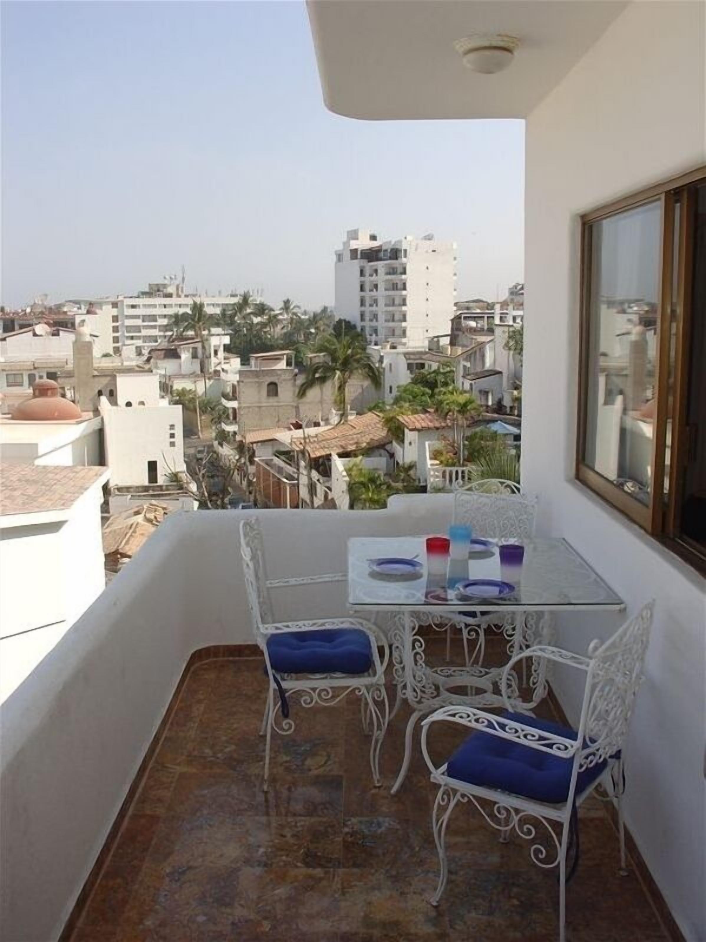 A balcony with a glass table and white metal chairs, overlooking a cityscape with various buildings and palm trees.