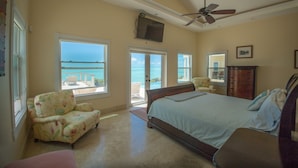 A bedroom with a large bed, a ceiling fan, a TV, and a view of the ocean through the windows.