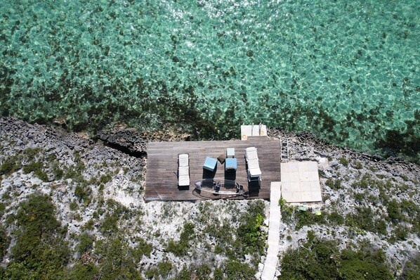 Vue aérienne d’une terrasse en bois avec des chaises longues et une petite table, entourée d’une eau turquoise cristalline et d’un paysage rocheux.