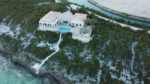 An aerial view of a coastal house with a swimming pool, surrounded by dense vegetation and a beach.