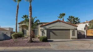 A single-story house with a stucco exterior, a tiled roof, and a garage. There are palm trees and a small garden in front.