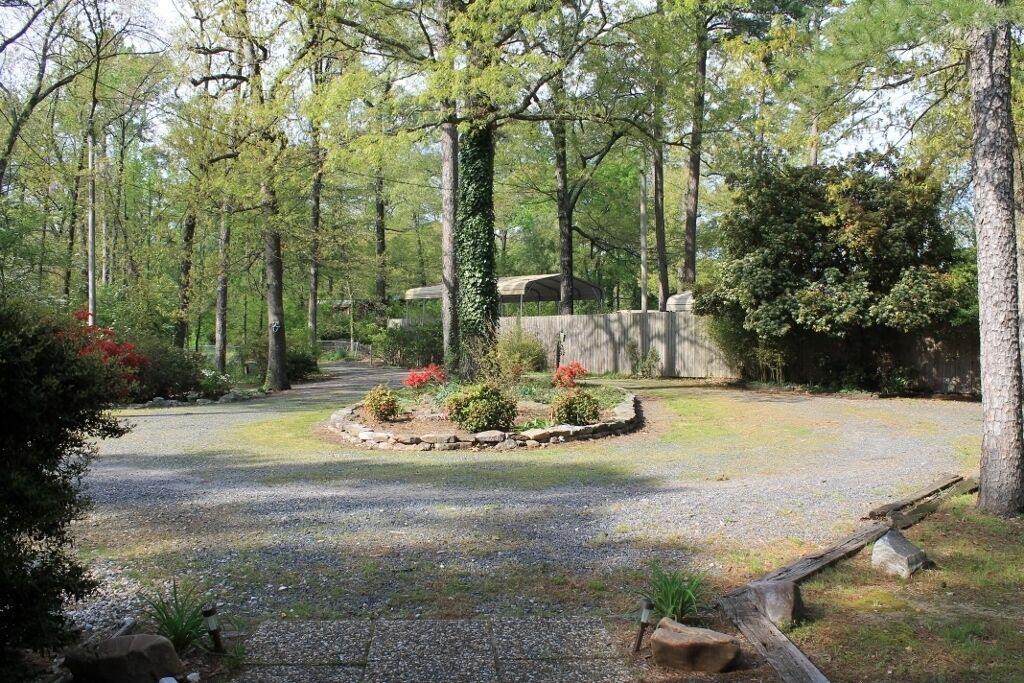 A forested area with a gravel path, a stone wall, and a covered structure in the background.