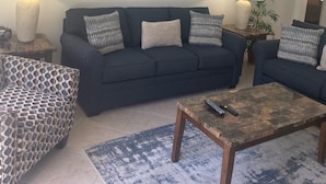 Una sala de estar con un sofá azul oscuro, un sillón estampado y una mesa de centro. Hay dos lámparas en mesitas auxiliares, un cuadro enmarcado en la pared y una planta en maceta.