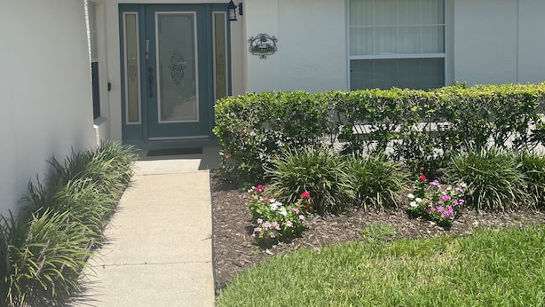Una casa de dos pisos con entrada principal, un pequeño porche y un jardín bien cuidado con diversas plantas y flores.