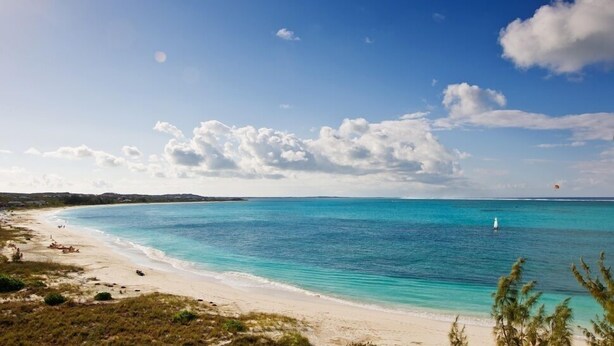 A beach with clear turquoise waters, a sandy shore, and a distant sailboat.