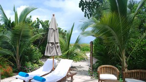 A sunlit outdoor seating area with lounge chairs and a wooden table, surrounded by lush greenery and a clear sky.