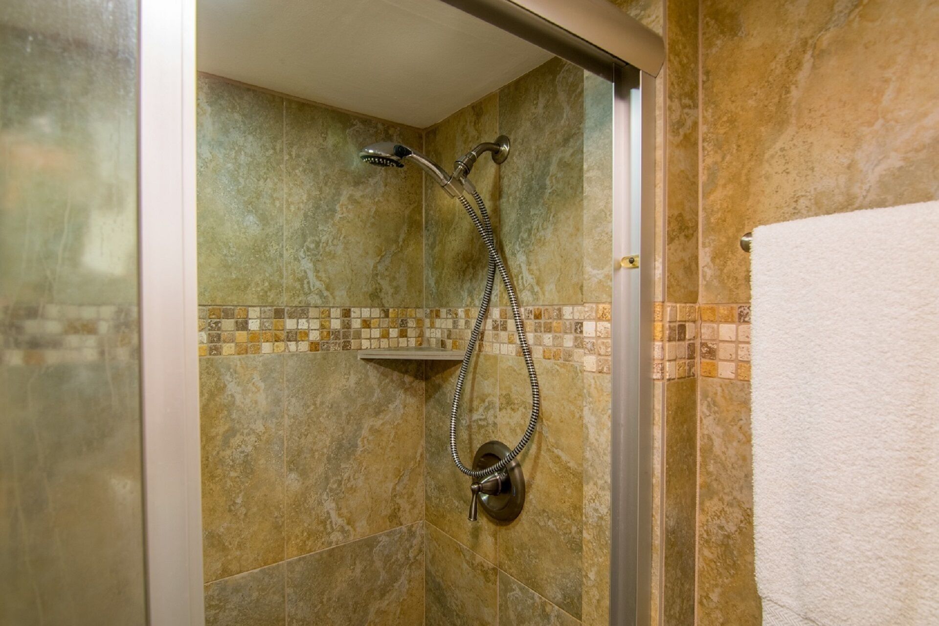 A shower with a handheld showerhead and a wall-mounted faucet, a tiled shower area, and a white towel hanging on the wall.