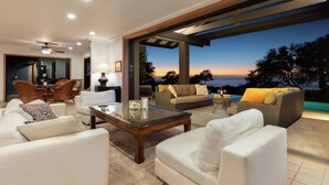 A modern living room with white sofas, a glass coffee table, and wicker furniture. It features a large window with a view of the ocean at sunset.