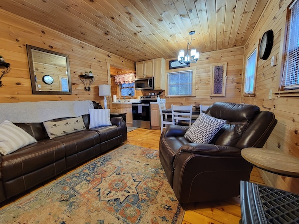 A cozy log cabin interior with a living area featuring a brown leather sofa and a leather recliner, a patterned rug, and wooden walls.