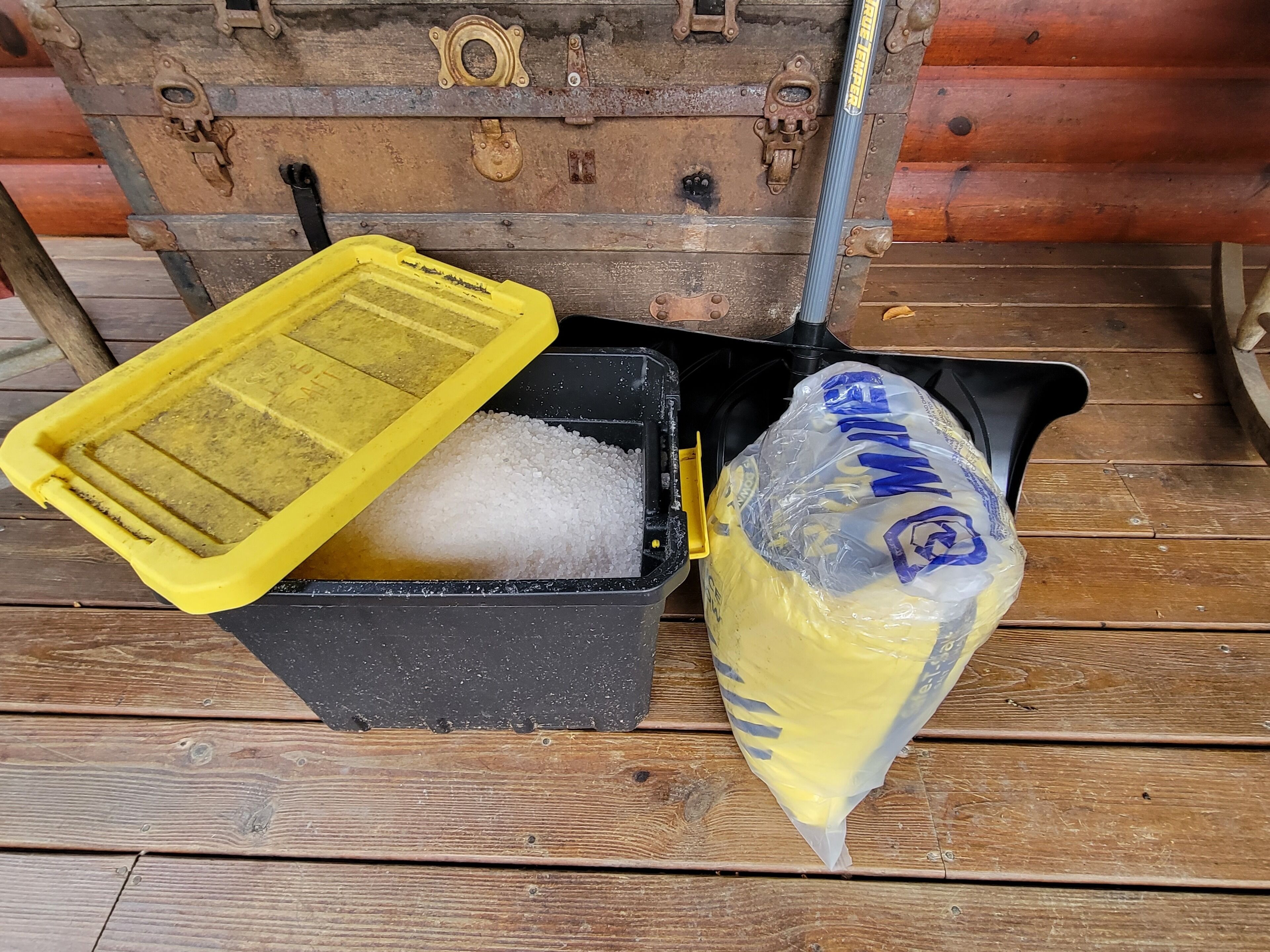 Un contenedor negro con tapa amarilla lleno de material granular blanco, junto a una bolsa de plástico con texto azul y etiqueta amarilla, sobre una cubierta de madera.