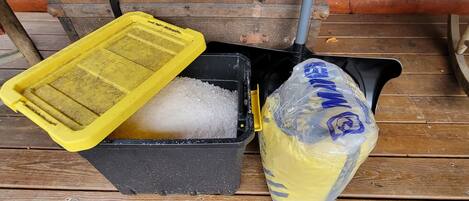 Un contenedor negro con tapa amarilla lleno de material granular blanco, junto a una bolsa de plástico con texto azul y etiqueta amarilla, sobre una cubierta de madera.