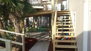 Eine Holzterrasse mit einer Treppe zu einem überdachten Bereich, umgeben von Palmen und einem grünen Tisch.