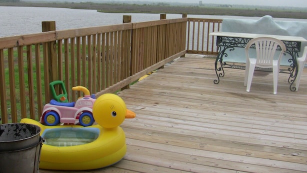 Eine Holzterrasse mit Blick auf ein Gewässer, ein gelber Schwimmdackel, ein blau-pinkes Spielzeugauto und ein weißer Stuhl.