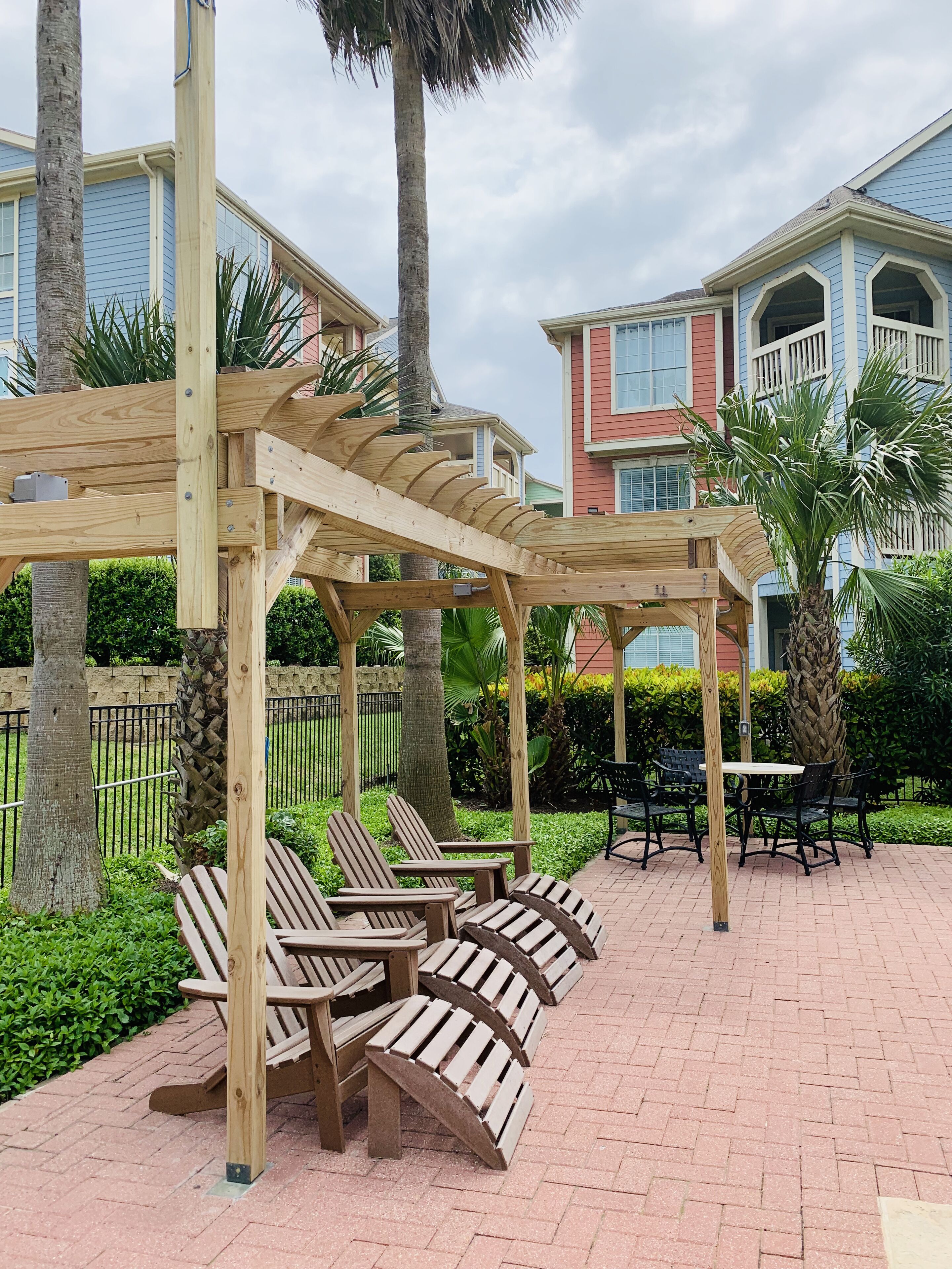 A wooden pergola with seating, surrounded by palm trees and a brick patio.