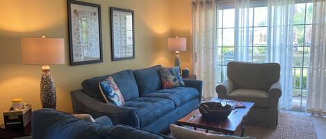 Una sala de estar con un sofá azul, un sillón beige, dos lámparas y una ventana con cortinas.