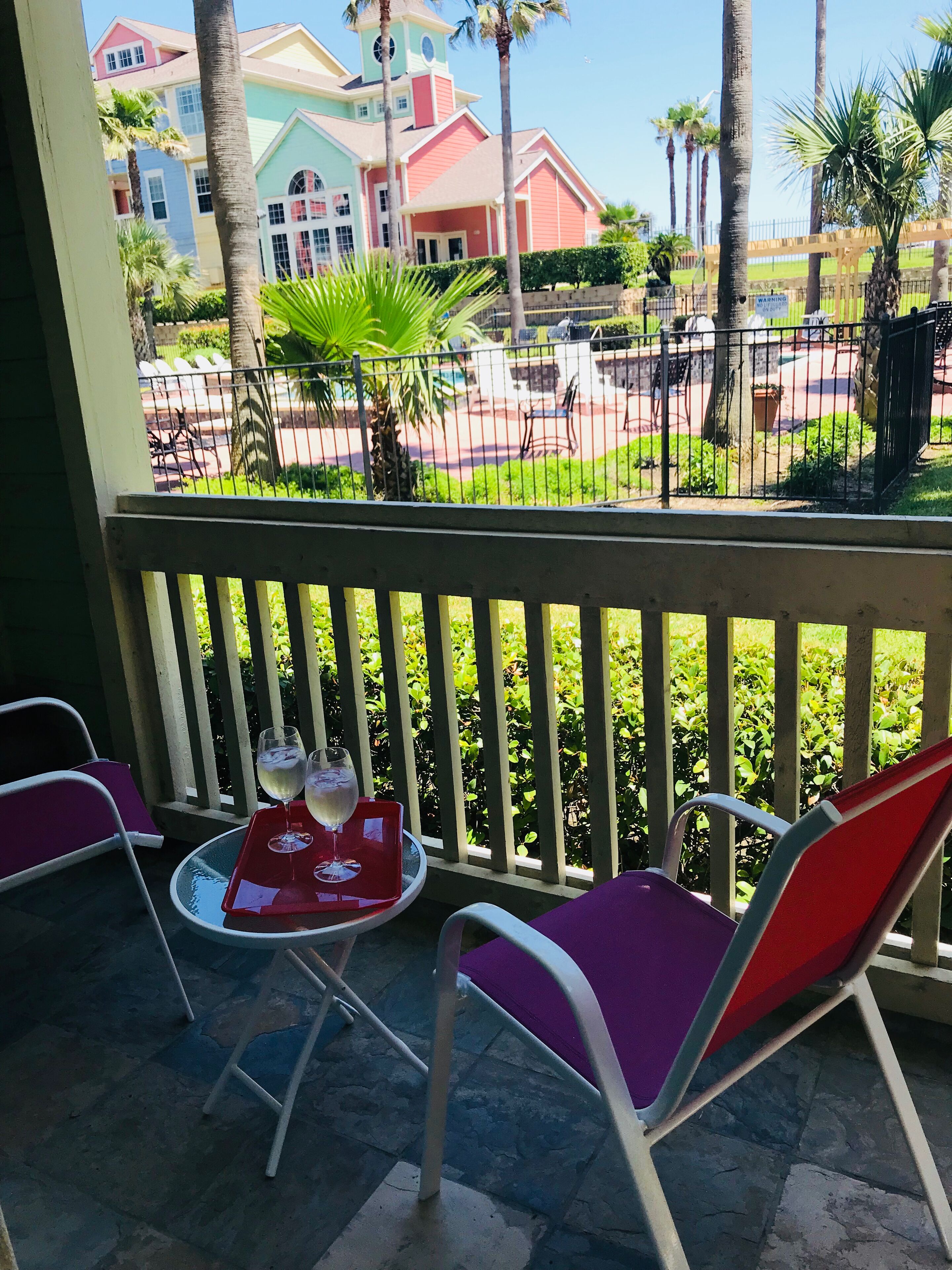 A balcony with two chairs and a table, a railing, and a view of colorful buildings and palm trees.