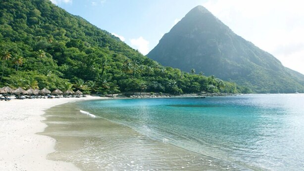 Une plage aux eaux turquoise et limpides, un rivage sablonneux et, en arrière-plan, une montagne verdoyante.