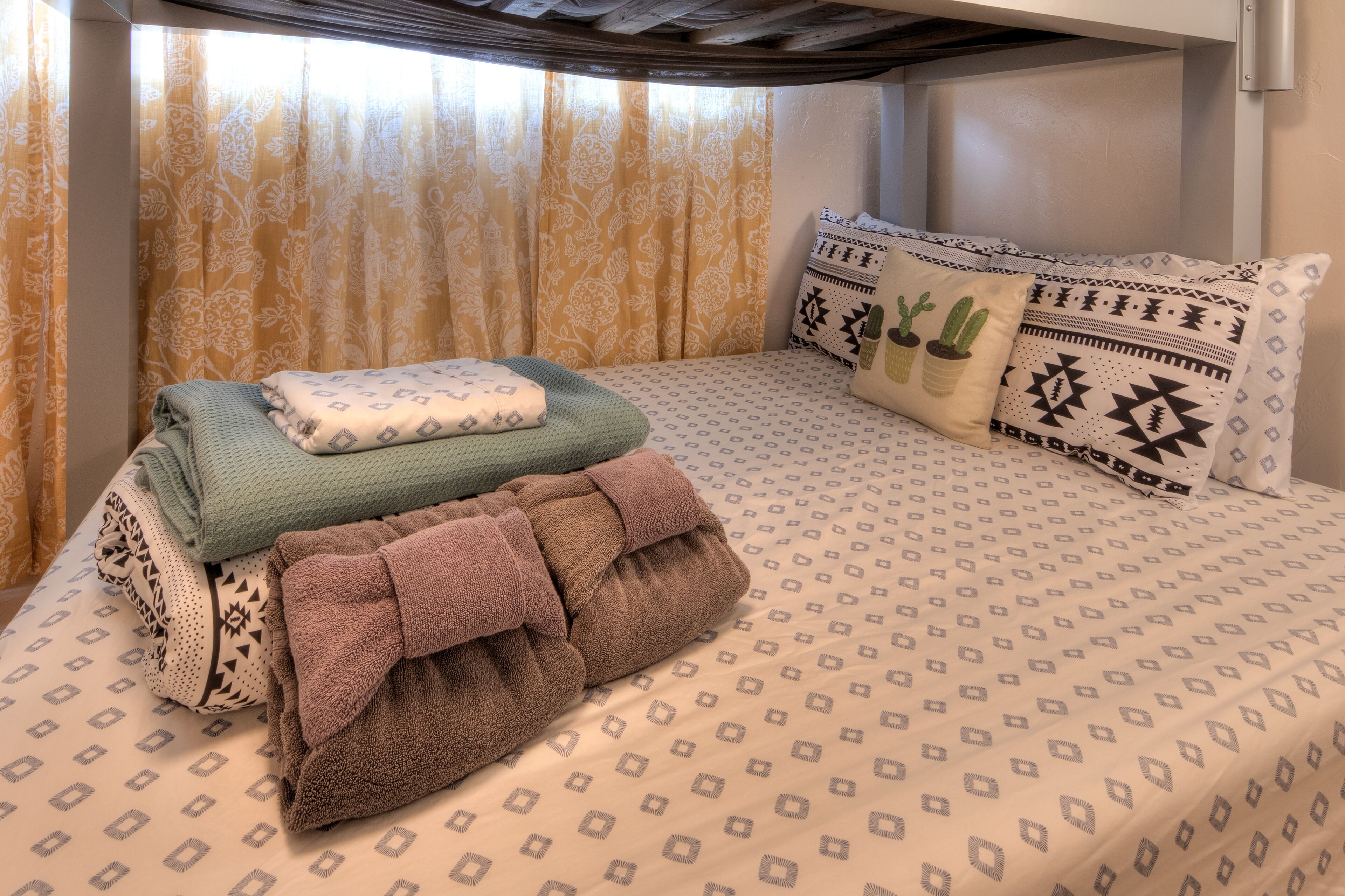 Una cama bien hecha con ropa de cama estampada, una toalla verde y marrón, y almohadones decorativos con diseños geométricos y motivos de cactus.