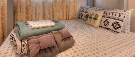 Una cama bien hecha con ropa de cama estampada, una toalla verde y marrón, y almohadones decorativos con diseños geométricos y motivos de cactus.