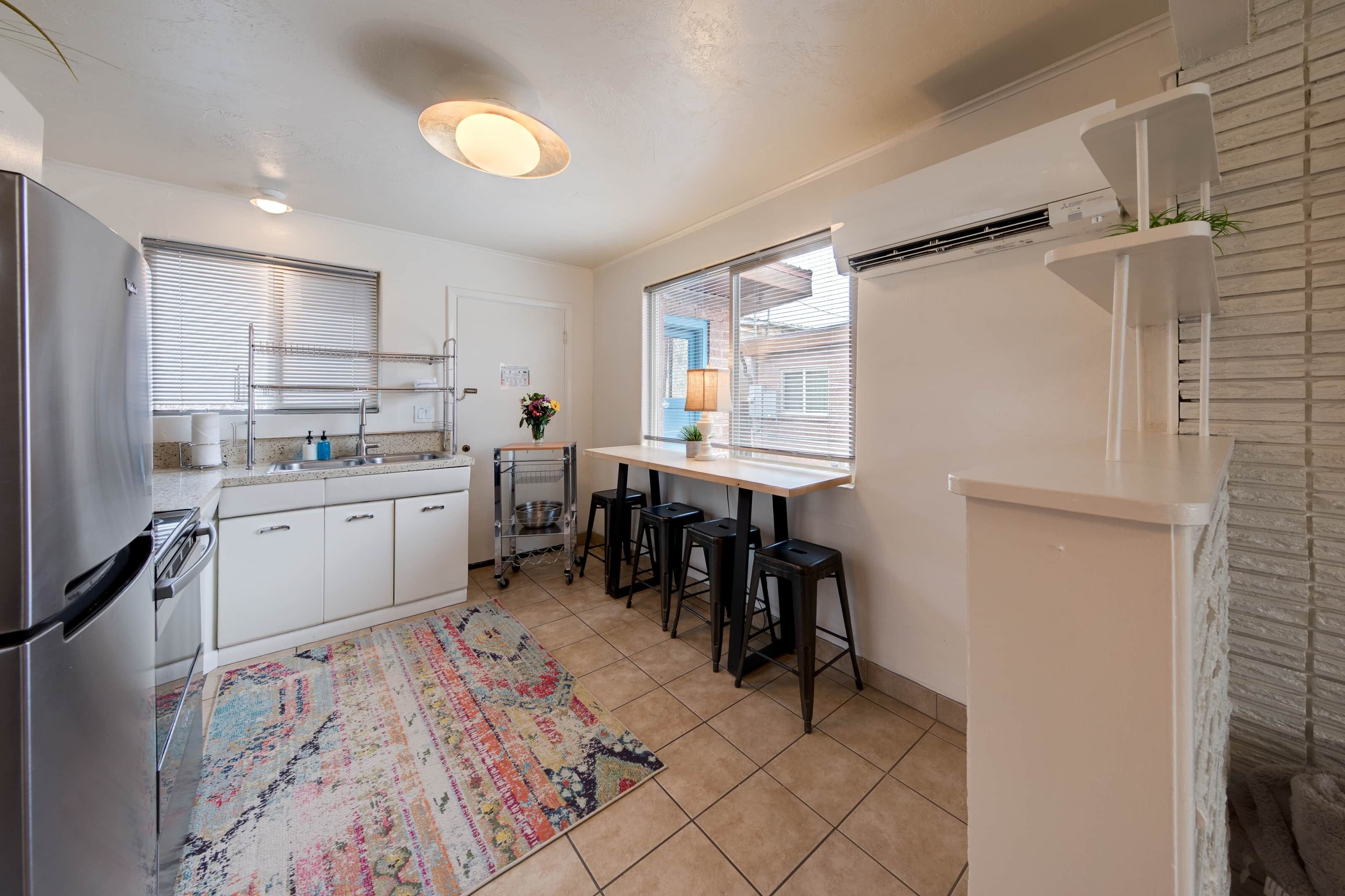 A compact kitchen with white cabinets, a stainless steel refrigerator, a small dining table with stools, and a patterned rug.
