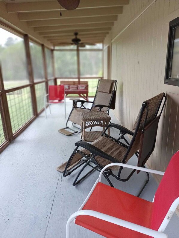 A covered porch with wicker chairs and a red chair, a table, and a ceiling fan.