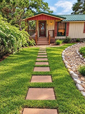 A house with a wooden porch, a stone pathway leading to the entrance, and a well-maintained lawn. - SERENE SMALL HOUSE ON POND WITH FOUNTAIN (Kennard)