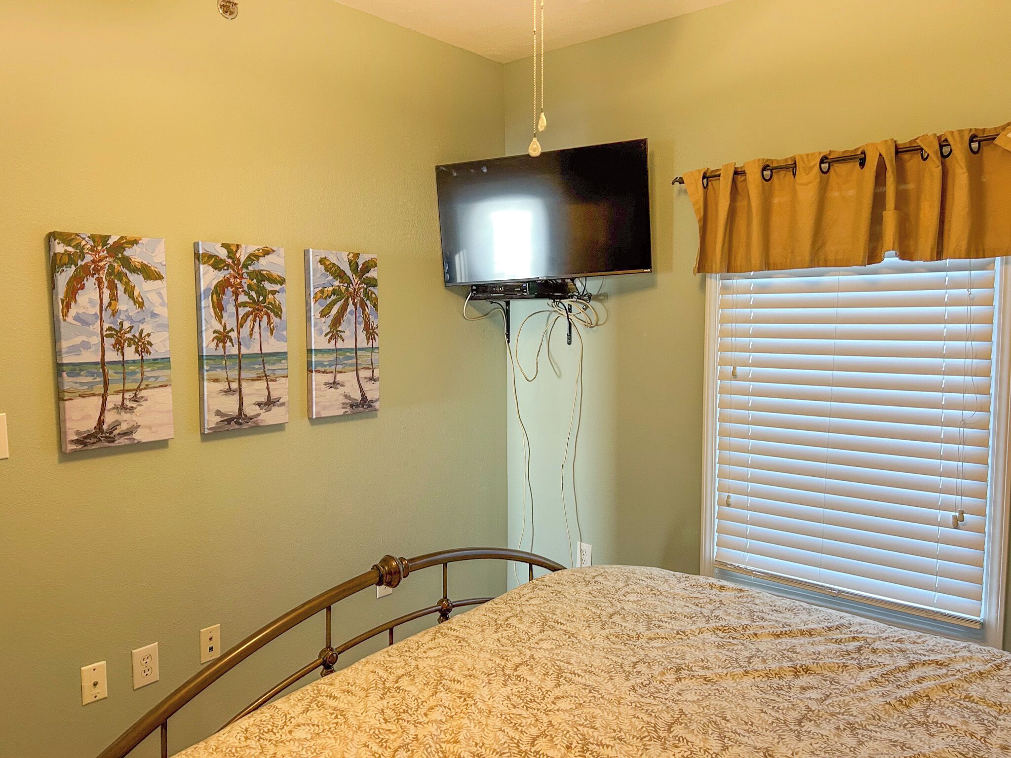 Quarto com uma cama, televisão fixada na parede e três quadros com tema praiano.