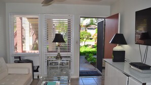 A living room with a white sofa, a glass coffee table with a book and remote, a lamp, and a TV on a stand. - Very,  very affordable. Steps to paved, oceanfront path. Oceanside, end unit (Kapaa)