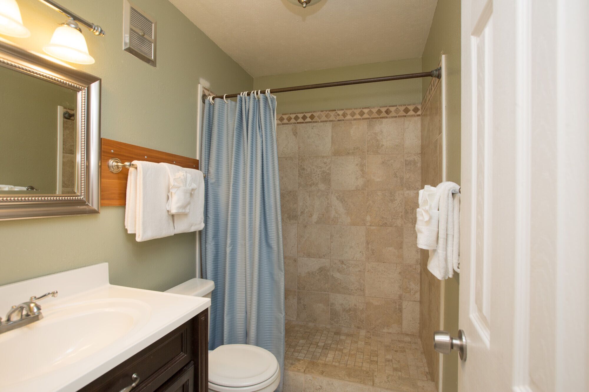 Un baño con un lavamanos blanco, un inodoro, una ducha con pared de azulejos color beige y una cortina de ducha azul.