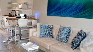 A modern living room with a sofa, patterned cushions, a glass coffee table, and a kitchen area with a dining table and chairs. - Very,  very affordable. Steps to paved, oceanfront path. Oceanside, end unit (Kapaa)