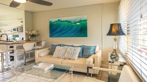 A modern living room with a beige sofa, a glass coffee table, and a painting of waves. - Very,  very affordable. Steps to paved, oceanfront path. Oceanside, end unit (Kapaa)