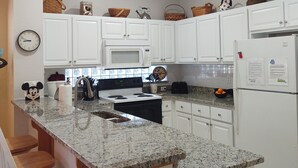 A kitchen with white cabinets, a granite countertop, a microwave, a stove, a refrigerator, and a sink. There are bar stools and a clock on the wall. - Kid Friendly Home W/Pool  in Gated Community,10 Min. to Disney (Kissimmee)