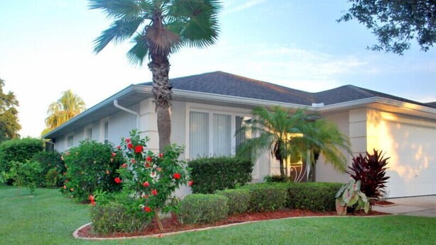 A single-story house with a well-maintained lawn, a palm tree, and a variety of shrubs and flowers. - Kid Friendly Home W/Pool  in Gated Community,10 Min. to Disney (Kissimmee)