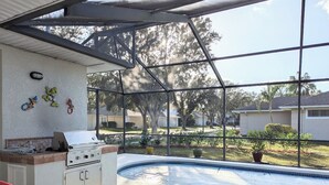 A covered outdoor swimming pool area with a grill, lounge chairs, and decorative wall art. - Kid Friendly Home W/Pool  in Gated Community,10 Min. to Disney (Kissimmee)