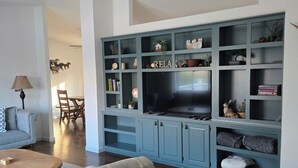 Un soggiorno con pavimento in legno scuro, un divano grigio con motivo decorativo, un tavolino rotondo, un televisore su un mobiletto blu, e un'area pranzo con tavolo e sedie in legno.