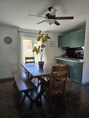 Un coin repas avec une table en bois, des bancs et des chaises. Un ventilateur de plafond est installé au-dessus, et il y a une fenêtre avec des rideaux.