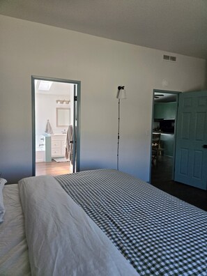 A bedroom with a bed, a bedside lamp, and an open door leading to a bathroom and a kitchen.